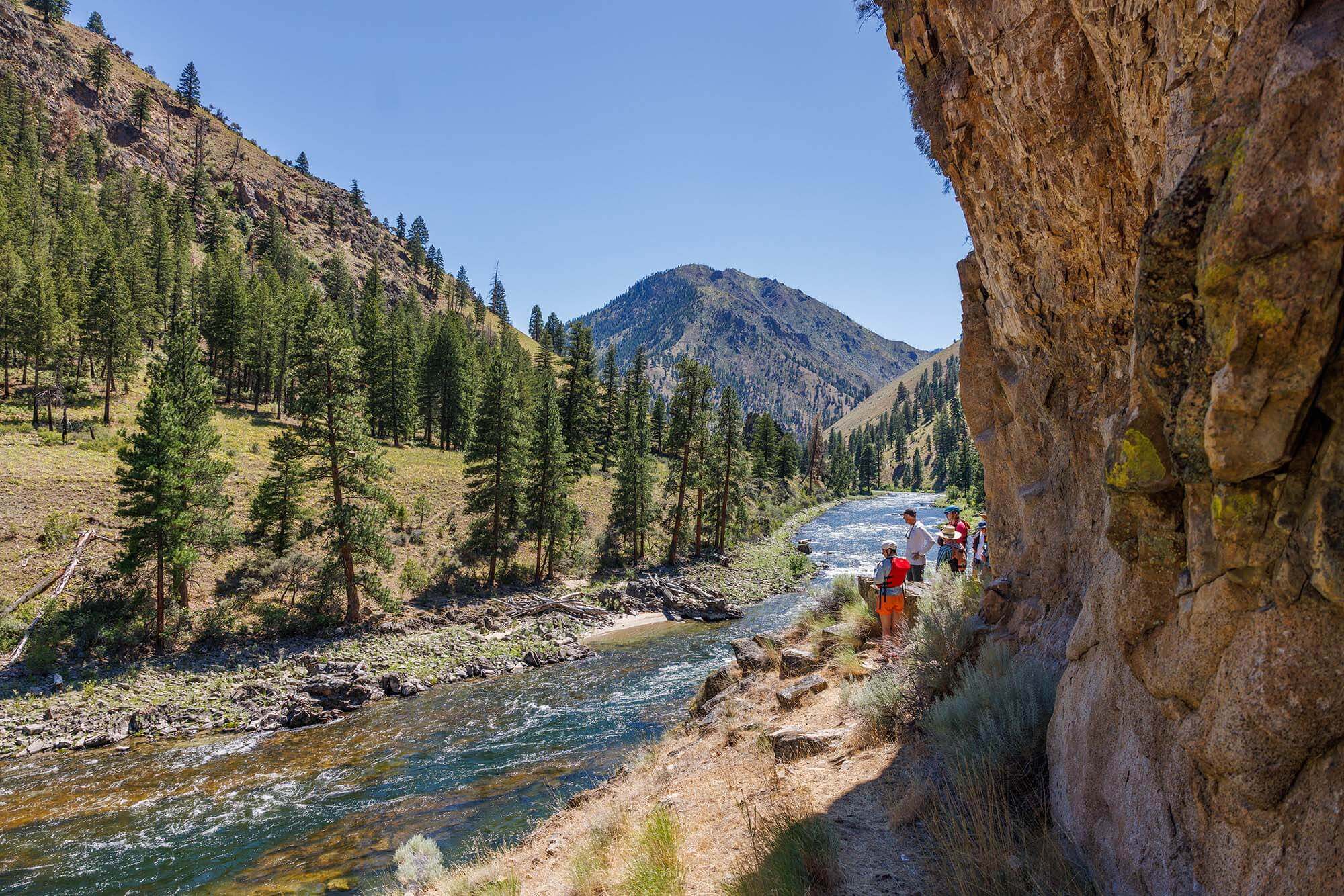 A group of people standing in front of a canyon wall at the edge of the Middle Fork of the Salmon River, and on the opposite side of the river, a tree-covered mountain.